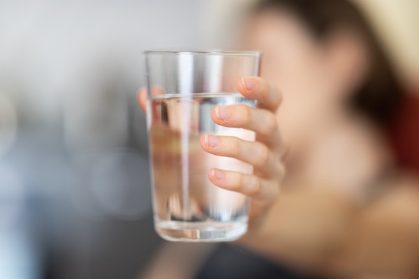 A women holding a glass of water. The glass of water is center of the image and focused, the woman is in the background and blurred.